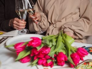 A couple toasting with champagne glasses over a bouquet of pink tulips on a dining table.