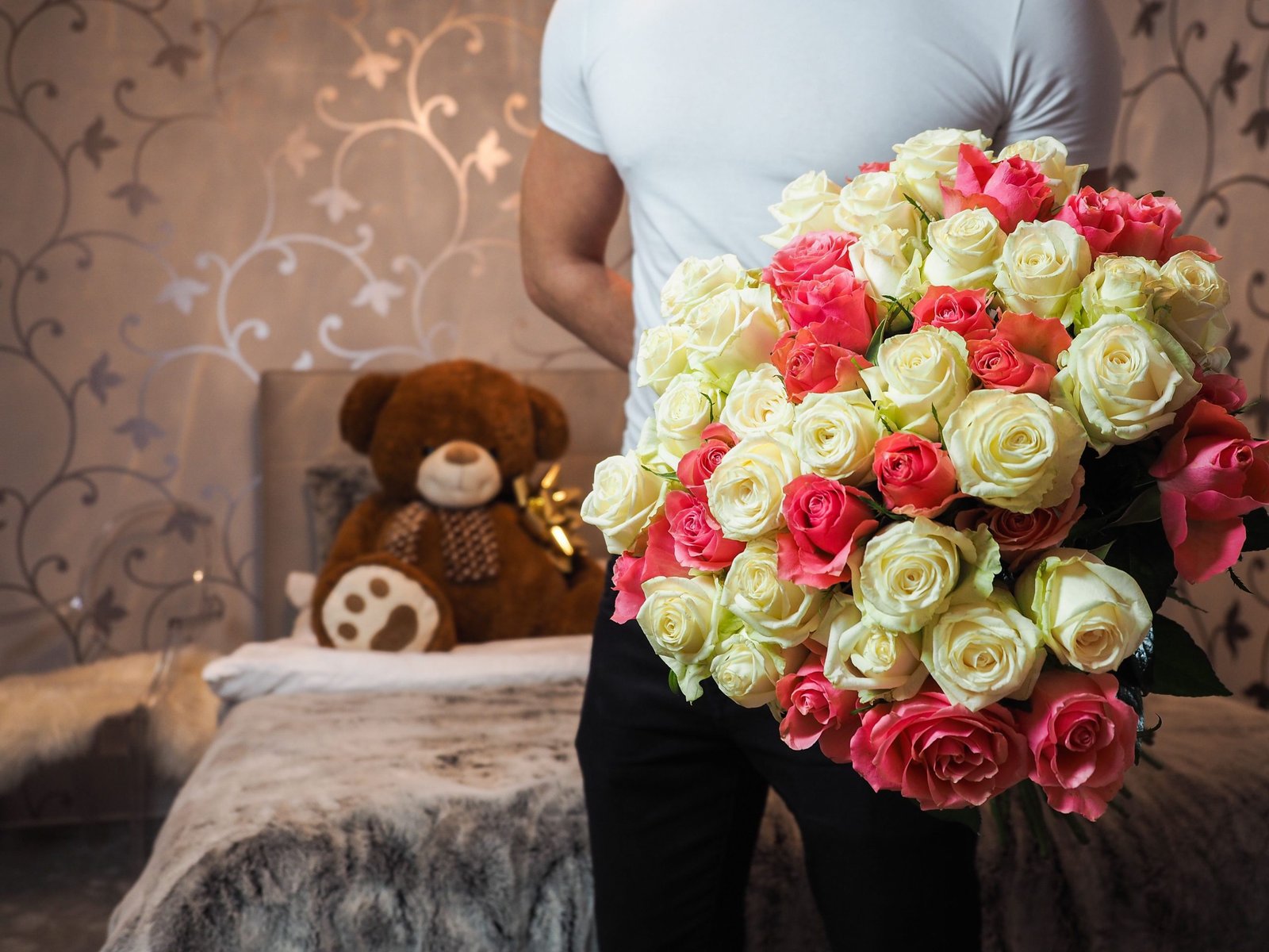 A man holding a bouquet of roses in a romantic indoor setting with a teddy bear.